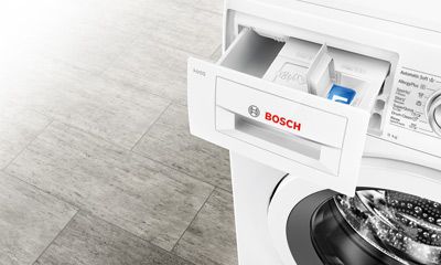 Das Bild zeigt eine digitale Steuerung für eine Waschmaschine, bei der eine Hand einen Finger auf ein Display mit verschiedenen Waschoptionen und Einstellungen legt. Das Display ist beleuchtet und zeigt Text und Symbole.